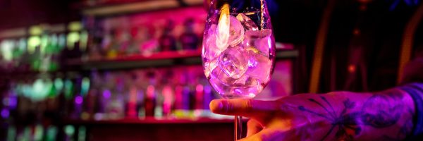 Close up of professional barman finishes preparation of alcoholic cocktail in multicolored neon light, gives it to client. Entertainment, drinks, service concept. Modern bar, trendy neoned colors.